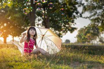 Cute young girl with beautiful colored umbrella sitting bubbles on green grass in the park at sunset time.Golden light.