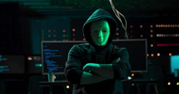 Dangerous Internationally Wanted Hacker with Masked Face Speaks into the Camera. In the Background His Operating Room with Multiple Displays and Cables.