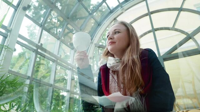 Young Pretty Woman With Long Blond Hair Female Wearing Blue Cloak Enjoying A Cup Of Coffee Sitting At Glass Table Near The Window In Cozy In Light Summer Cafe Low Angle Shot