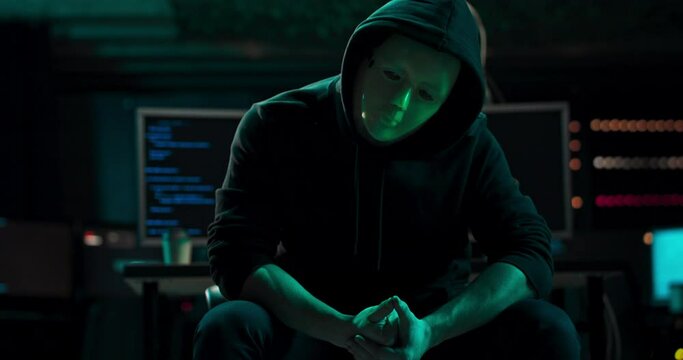 Wanted Hacker with Masked Face looks into the Camera. In the Background His Operating Room with Multiple Displays and Cables. After a while he turns away from the camera and goes back to work.