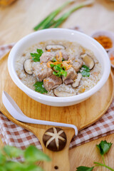 Pork boiled rice with shiitake mushrooms on a white bowl with a spoon placed on a wooden table for breakfast.