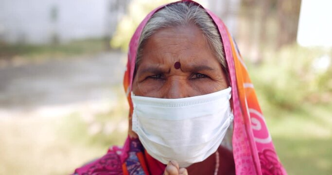 Handheld slow-motion portrait of an elderly women speaks to looking at camera pov wearing face mask to request in prayer position joining hands for Namaste with sign gestures, outdoors