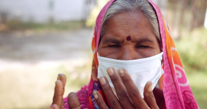 Handheld slow-motion portrait of an elderly women speaks to looking at camera pov wearing face mask to request in prayer position joining hands for Namaste with sign gestures, outdoors