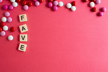 the word fave written as a flat lay in wood scrabble tiles on a plain red background surrounded by red, white, and pink candies