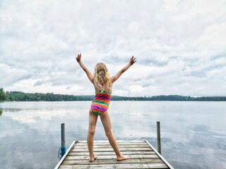 Blonde girl in swimsuit standing on wooden lake river dock with her hands raised up. Harmony with nature and happy healthy active childhood. Summer outdoor water activity. View from back behind.