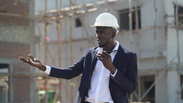 Angry african american architect in suit and hard hat talking by walkie talkie control and communication to blame worker outdoors on construction site. contractor using radio operation in industry