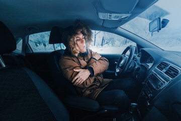 Fototapeta Man in down jacket tries to keep warm and not freeze in his stalled car in winter. The concept of a dangerous accident or heater failure in the cold season obraz