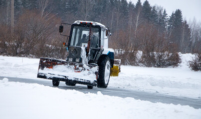 an old snow-covered tractor with  blade removes snow from the road, sweeps snow from the road with  large brush, in  severe blizzard.