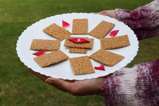 Female hand holding Winter special sweet dish of White sesame and jaggery melted in clarified butter and rolled in to sheets garnished and decorated with fresh rose petals on white plate