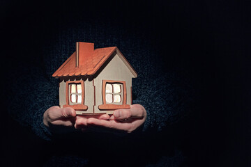model of a wooden house in hands on a dark background, bright light in the windows, the concept of a warm, cozy home, real estate agency, insurance and mortgage