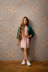 Fototapeta na wymiar beautiful child girl with long beautiful hair in a pink dress and a bright leather jacket against the background of a wall with wallpaper