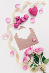Valentine's Day composition. Blank greeting card mockup in envelope, rose, macaroon, gold ribbon, heart and rose petals. White painted wood background. Template for your design. Top view, flat lay.