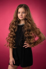 Fototapeta premium beautiful girl child with long well-groomed hair in a beautiful hairstyle in a black overalls on a bright pink background