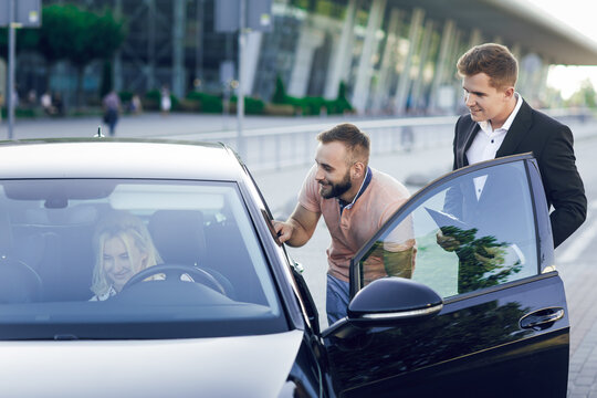 A young car dealer in a business suit shows buyers a new car. Young couple, man and woman, buy a car. Woman sitting behind the wheel. Purchase of machines, test drive