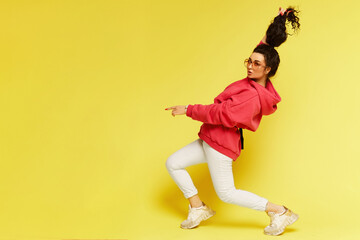 Fototapeta na wymiar Young stylish brunette woman in pink hoodie pointing finger at the left side of the shot, isolated with copy space. Model girl in summer hipster sport clothes posing on the yellow background isolated