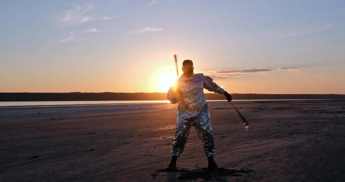 A guy in a metal suit with his hands rotates fiery torches on chains, against the backdrop of a sunset in the middle of a large beach. Master plan.