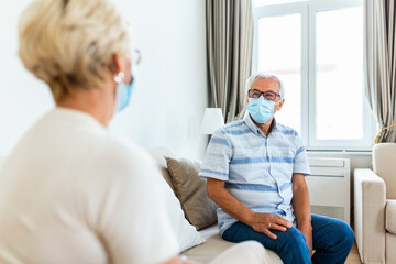 Old friends separated by social distancing on sofa at home. Senior friends wearing protective mask, preventing covid 19 coronavirus pandemic infection spread. Social distancing on sofa at home