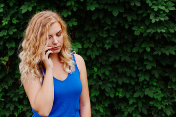 Naklejka premium Beautiful young girl in a blue dress talking on the phone. Against the background of wild grapes, summer day. Copy space.