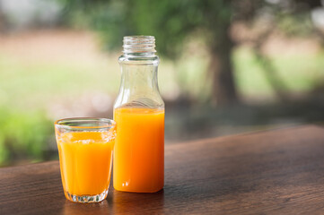 Fresh orange juice in no label clear plastic bottle in small glass on wooden table, natural trees blur in the background. With copy space.