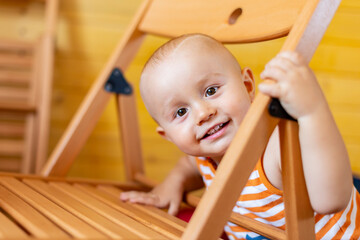 A portrait of a cute adorable laughing smiling baby boy of 12 months or 1 year old looking out from behind a chair wearing a sleeveless striped orange top.