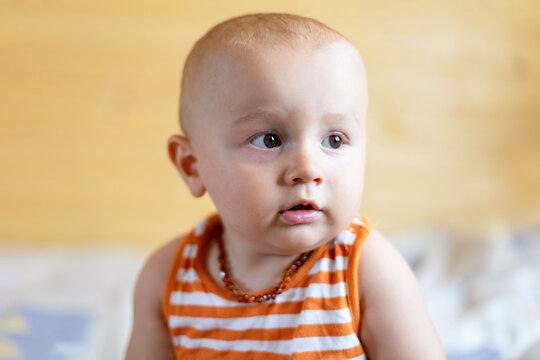 A portrait of a cute adorable curious  baby boy of 12 months or 1 year old looking aside wearing amber teething baby necklace in a sleeveless striped orange top.