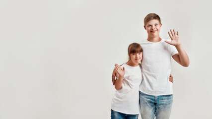 Two kids, disabled boy and girl with Down syndrome smiling and waving at camera, embracing each other while posing together isolated over white background