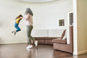 Full length shot of loving grandmother playing with her adorable little granddaughter, lifting her up in the air while having fun together in a living room at home