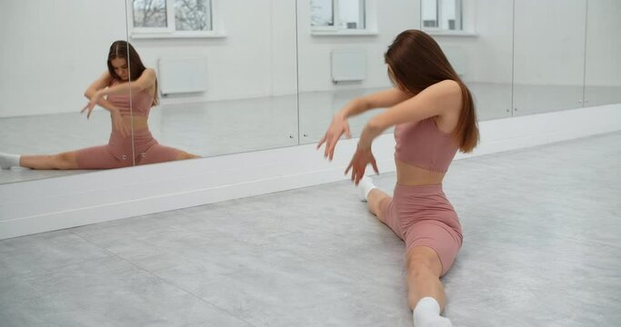 Dancer is sitting on the twine and warming up infront of the mirror wall in the white bright dance hall, ballet rehearsal, ballerina in the ballet studio, dance hall lesson, 4k 60p Prores HQ