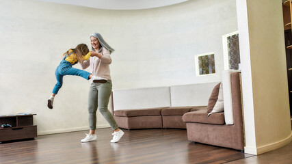 Fototapeta na wymiar Full length shot of loving grandmother playing with her adorable little granddaughter, lifting her up in the air while having fun together in a living room at home