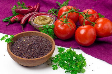 Black mustard seeds in wooden bowl and fresh vegetables. Mustard grain, tomatoes, red chili pepper, parsley and mustard sauce on table cloth. Organic, healthy food