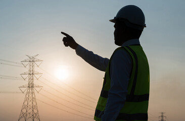 An Asian male engineer works in front of a high-voltage tower pointing the finger. In the sunset the sky is beautiful