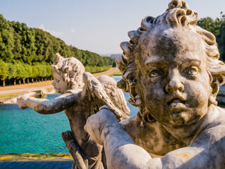 Detail of the fountain of Venus and Adonis with the face of a winged marble cherub in the foreground, Royal Palace of Caserta, Italy
 - obrazy, fototapety, plakaty