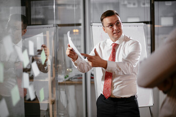 Business people in conference room use sticky notes on glass wall. Colleagues working on new project.