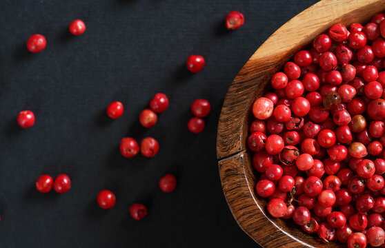 Whole Pink Or Red Peppercorns In Small Wooden Bowl, Some Scattered On Black Gray Paper, Closeup View From Above