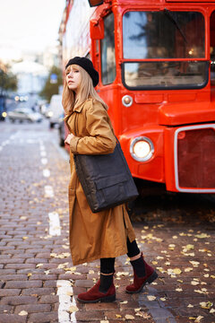 Blonde young woman in black beret and orange trench on the city street with doubledecker background. Female looking forward at the street. Romantic look image