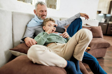 Spending time with grandson at home. Cute little boy playing video game on smartphone while relaxing on sofa with his happy grandfather