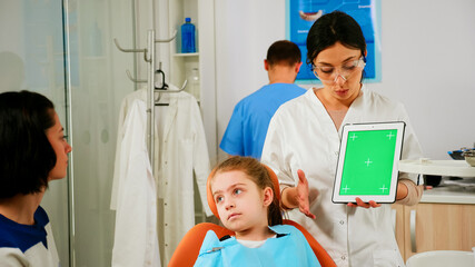 Stomatologist woman looking at green screen tablet while speaking with mother of little patient. Pediatric dentist explaining using monitor with green chroma key izolated chroma pc key mockup display