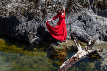 A blonde woman in a long red dress in a mystical image soars in the air above the green water of the lake against the background of a stone shore in the Altai mountains. Magic and levitation.