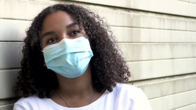 Beautiful mixed race African American girl biracial teenager young woman outside wearing a face mask and using her mobile cell phone during COVID-19 Coronavirus pandemic