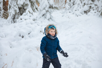 Fototapeta na wymiar boy in a ski suit and ski goggles stands on a ski slope. Child playing with snow in the forest