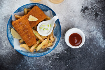Above view of fish and chips with dips served in a blue bowl, horizontal shot on a dark grey stone background with space