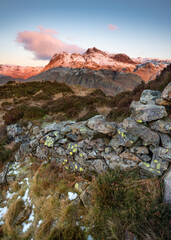 Fototapeta na wymiar Old rural stone wall leading to view of snowcapped Cumbrian mountains; The Langdale Pikes as morning sunlight casts a pink glow. Lake District, UK.