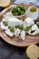 Feta cheese with giant green olives, olive oil and lemon on a round wooden tray, vertical shot, close-up