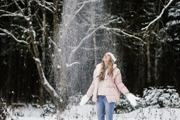 Fototapeta na wymiar beautiful girl in a pink jacket and white hat, laughs throws up snow and stands in a snowy cloud in the forest