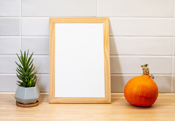 Obraz premium Mock up poster on coutertop table in white kitchen with green plant and a pumpkin. Empty frame for picture background. Interior design