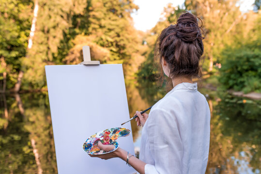 woman paints picture landscape, girl artist, view back, summer park lake pond river, white shirt , creating creativity artistic mood. Blank white canvas, getting started. Brush color palette paints.