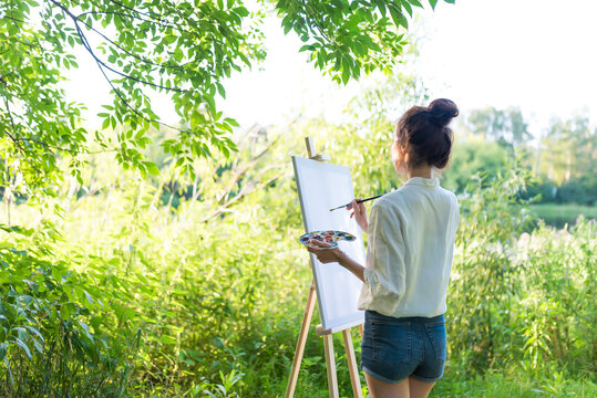 Free space copy text, woman draws picture landscape, summer pond lake, girl artist, white shirt, creating creative artistic. Blank white canvas, getting started. Brush and color palette with paints.