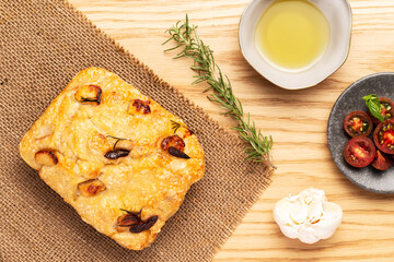 Fototapeta na wymiar Italian focaccia with confit garlic cloves and rosemary, alongside tomatoes, garlic bulb, olive oil and rosemary twig on a jute cloth lying on a wooden table. Top view.