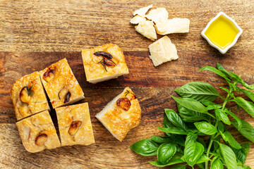 Italian focaccia cut in pieces with garlic cloves and rosemary, alongside some olive oil, parmesan cheese, and basil on a wood chopping board. Top view.