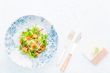 Smoked salmon salad with Arugula, apple, enoki mushrooms, zucchini and beetroot on a white and blue plate aside silver cutlery and a quenelle of cheese with dill over a blue background with copy space